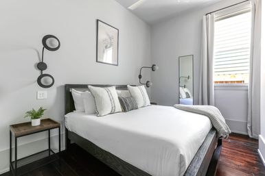 Bright minimalist apartment bedroom with a queen bed dressed in crisp white linens and striped pillows, dark wood frame and floors, gray walls, round black wall sconces, small wooden nightstand with potted plant, full-length mirror and curtained window letting in natural light.