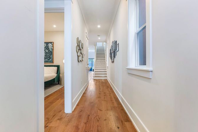 Bright modern residential hallway with warm hardwood floors, white walls, gallery picture frames and geometric mirrors, open bedroom doorway to the left, and a staircase leading to the upper level.