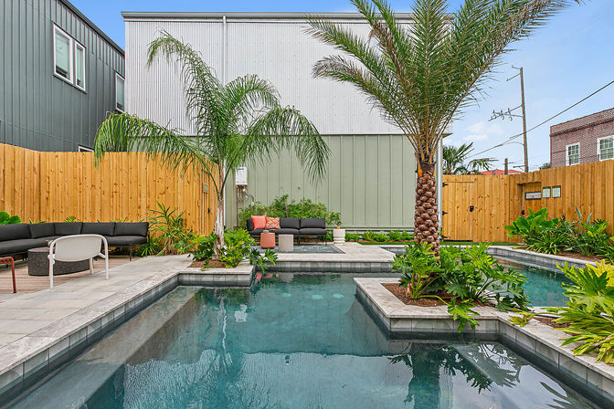 Modern urban backyard courtyard with a rectangular pool, palm trees and tropical plants, wooden privacy fence, gray-siding building backdrop and contemporary outdoor lounge seating on stone pavers.