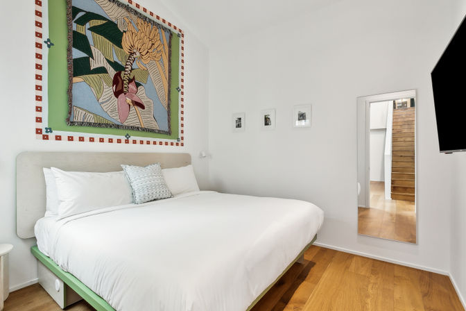 Bright, cozy minimalist bedroom with a king-size bed dressed in white linens, patterned accent pillow, colorful tropical tapestry above the headboard, three small framed photos, full-length mirror reflecting a wooden staircase, and warm hardwood floors.