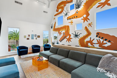 Bright modern living room with vaulted ceiling, bold orange crocodile mural, teal sectional sofa, blue club chairs, orange acrylic coffee table, windows showing palm and blue sky.