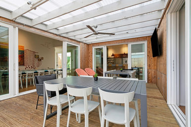 Modern covered patio with wooden deck and translucent roof, gray outdoor dining table and white chairs, ceiling fan, sliding glass doors to indoor dining and kitchen, pink lounge chairs and wall-mounted TV.