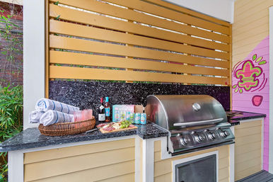 Outdoor patio kitchen with stainless steel gas grill, black granite countertop, yellow slatted privacy screen, wicker basket of rolled towels and glasses, charcuterie board with wine and canned drinks, colorful pink mural.