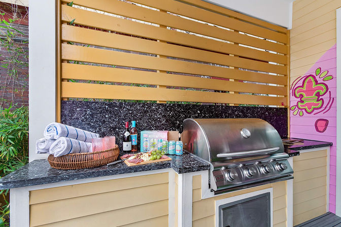 Outdoor patio kitchen with stainless steel gas grill, black granite countertop, yellow slatted privacy screen, wicker basket of rolled towels and glasses, charcuterie board with wine and canned drinks, colorful pink mural.