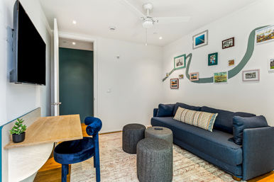Modern apartment living room with navy sofa and patterned lumbar pillow, gallery wall with teal accent stripe, round gray ottomans, wall-mounted TV, wooden desk and blue velvet chair.