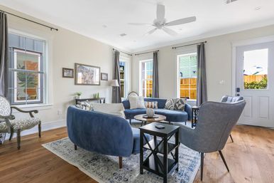 Bright modern living room with curved blue velvet sofa and gray armchairs forming a cozy conversation circle around a round coffee table on a patterned rug, hardwood floors, tall windows with gray curtains, white ceiling fan, and a door to the yard.