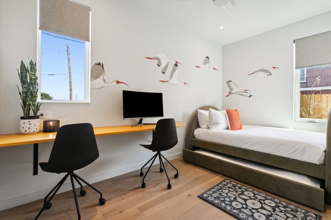 Bright modern guest bedroom and home office with a yellow wall-mounted desk, two black swivel chairs and monitor, twin daybed with trundle, flying bird wall decals, potted plant, and hardwood floors.