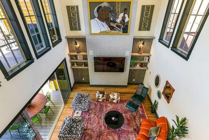 Vibrant eclectic two-story loft living room with tall windows, hardwood floors, wall-mounted TV beneath large artwork, colorful area rug, mixed seating and sliding patio door to balcony.