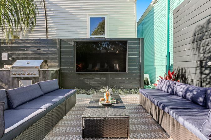 Sunny urban backyard patio with two navy-cushioned wicker sofas facing a glass-topped rattan coffee table, wall-mounted flat-screen TV on a black fence, stainless-steel grill, patterned outdoor rug and potted plants