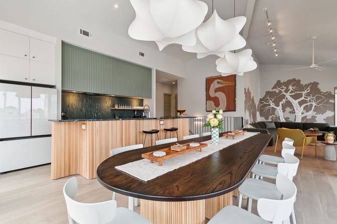 Open-plan modern dining room and kitchen with a long dark wood table and white chairs, fluted light‑wood island and bar stools, green tiled backsplash, oversized white sculptural pendant lights, tree mural and cozy seating area