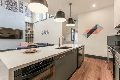 Modern urban loft-style open-concept kitchen with white quartz island, built-in sink and dishwasher, matte black pendant lights, stainless appliances, hardwood floors, high ceilings, and geometric wall art.