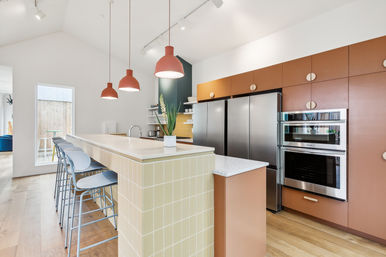 Bright modern open-plan kitchen with yellow-tiled island and breakfast bar, metal bar stools, stainless-steel appliances, terracotta cabinets, pendant lights and hardwood floors.