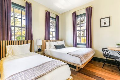 Bright, cozy modern guest room with two low wooden platform beds (twin and double), tall black-framed windows with purple curtains, bedside lamp, small desk and chair, and warm hardwood floors