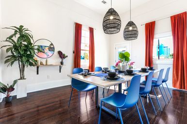 Vibrant sunlit contemporary dining room with a long wooden table set for eight, bold blue chairs, woven black pendant lights, rust-red curtains, large windows with city view, and lush potted plants on dark hardwood floors.