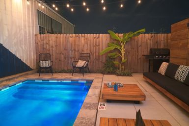 Cozy backyard patio at night with a small illuminated blue pool, string lights overhead, wooden fence, two rattan chairs with patterned pillows, a banana plant, low wooden coffee table, cushioned outdoor sofa and a barbecue grill.