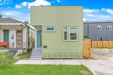Compact pastel-green modern rowhouse with horizontal siding, small front stoop and two front windows, fenced yard and neighboring homes on an urban residential lot under a bright blue sky.