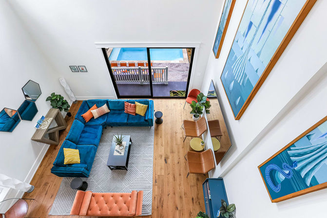 Aerial view of a modern two-story living room with a blue velvet L-shaped sofa, orange tufted bench, hardwood floors, large blue abstract wall art and sliding glass doors showing a backyard pool.