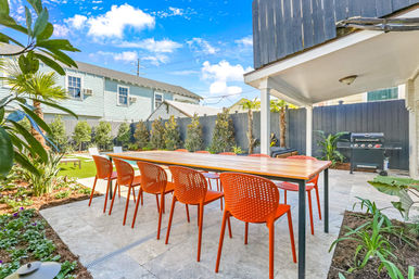 Sunny residential backyard patio outdoor dining area with long wooden table and eight bright orange chairs on stone tile, black privacy fence, gas grill, tropical plants and small lawn