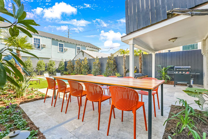 Sunny residential backyard patio outdoor dining area with long wooden table and eight bright orange chairs on stone tile, black privacy fence, gas grill, tropical plants and small lawn
