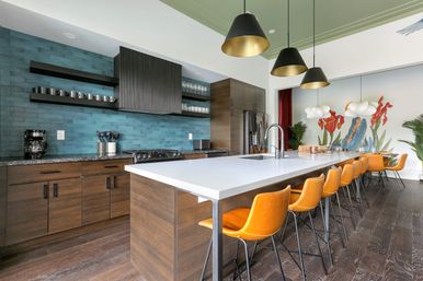 Modern open-concept kitchen with a long white island, teal subway tile backsplash, dark wood cabinets, orange leather bar stools, black pendant lights, and hardwood floors