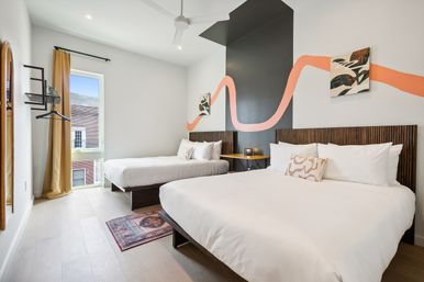 Bright urban hotel-style bedroom with two large white beds on low wooden platforms, dark slatted headboards, black accent wall with a peach ribbon mural, bedside table and lamp, ceiling fan, tall window with mustard curtain and light wood floors.