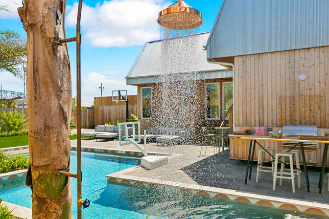 Sunny suburban backyard pool scene with a copper outdoor shower cascading into a turquoise pool, wood-clad house, poolside bar with stools, lounge seating and basketball hoop.
