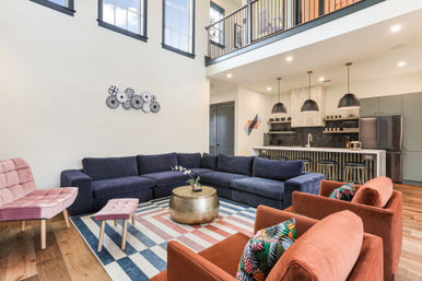 Bright modern open-concept living room and kitchen with a navy L-shaped sectional, rust armchairs, pink accent chair, striped area rug, brass round coffee table, pendant lights, and a second-floor balcony overlooking the space.