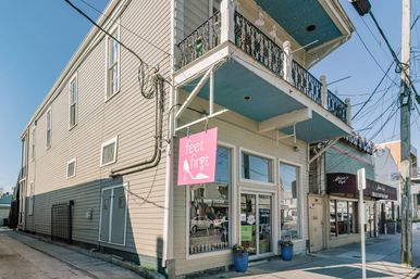 Sunny small-town corner storefront in a beige two-story building with a pink hanging sign, glass entry flanked by blue planters, ornate second-floor balcony and overhead utility wires.