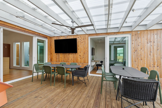 Bright covered patio with wooden walls and deck, two outdoor dining tables with green chairs and wicker seats, ceiling fan, wall-mounted TV and sliding glass doors to the interior.