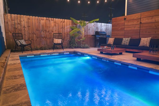 Modern backyard swimming pool glowing blue at night with tiled patio, wooden fence, string lights, outdoor sofa and cushions, low coffee tables, wicker chairs and a gas grill.