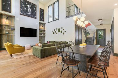 Bright open-concept living and dining area with two-story ceiling, olive green sectional, mustard accent chairs, wall-mounted TV, large wooden dining table with black metal chairs, wood floors, staircase mural and globe pendant lights.