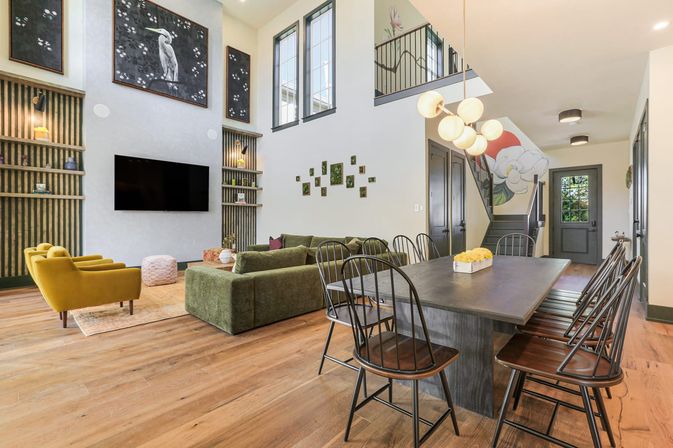 Bright open-concept living and dining area with two-story ceiling, olive green sectional, mustard accent chairs, wall-mounted TV, large wooden dining table with black metal chairs, wood floors, staircase mural and globe pendant lights.