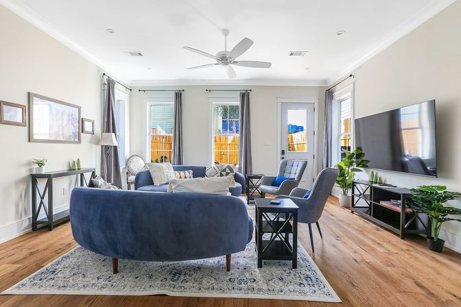 Bright contemporary living room with navy curved velvet sofa, gray accent chairs, hardwood floors, patterned rug, wall-mounted TV, potted plants, and sunlit windows and door.