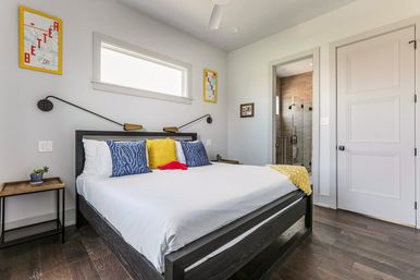 Bright modern bedroom with dark wood platform bed, white linens and blue, yellow and red accent pillows, wall-mounted swing lamps, small horizontal window, hardwood floors and glass-enclosed en-suite shower.