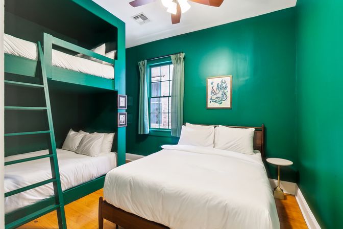 Cozy emerald-green bedroom in a vacation rental with built-in bunk beds and a double bed dressed in crisp white linens, hardwood floors, a window with green curtains, ceiling fan, and framed artwork.