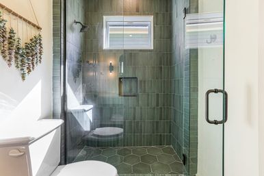 Bright modern bathroom with glass-enclosed shower featuring vertical green subway tiles, hexagon green floor tiles, small window, white toilet and hanging eucalyptus wall decor.