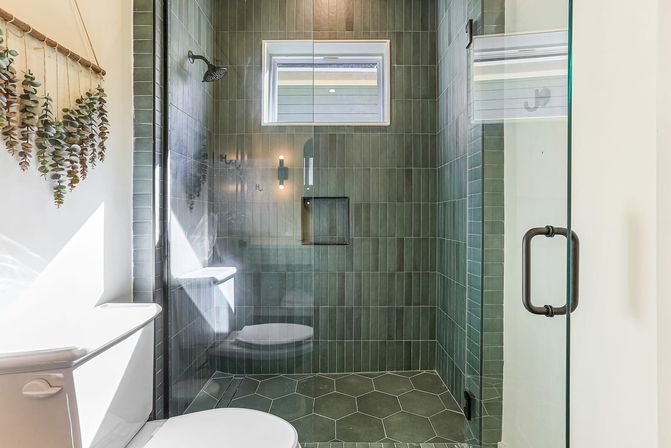 Bright modern bathroom with glass-enclosed shower featuring vertical green subway tiles, hexagon green floor tiles, small window, white toilet and hanging eucalyptus wall decor.