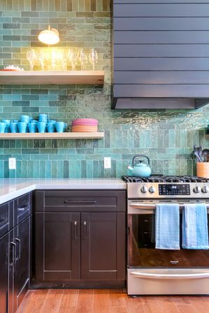 Modern kitchen corner with glossy green subway tile backsplash, dark wood cabinets, stainless steel gas range with teal kettle, open wood shelves of blue mugs and colorful plates, wine glasses lit by a wall sconce and warm hardwood floor.