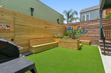 Sunny urban backyard courtyard with bright artificial turf, built-in wooden benches and raised planter, colorful mural on a wooden fence, palm tree, exterior stairs and a grill.