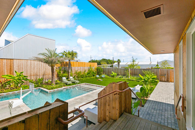 Sunny backyard oasis with covered porch and wooden deck steps leading to a rectangular swimming pool, tiled patio, lounge chairs, palm trees and a fenced grassy yard.