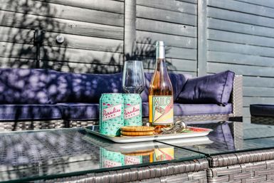 Sunlit backyard patio with a glass-top wicker coffee table holding a bottle of rosé, two pastel canned drinks, an empty wine glass and snacks on a white tray, in front of a purple-cushioned outdoor sofa and grey wood fence — ready for al fresco sips.