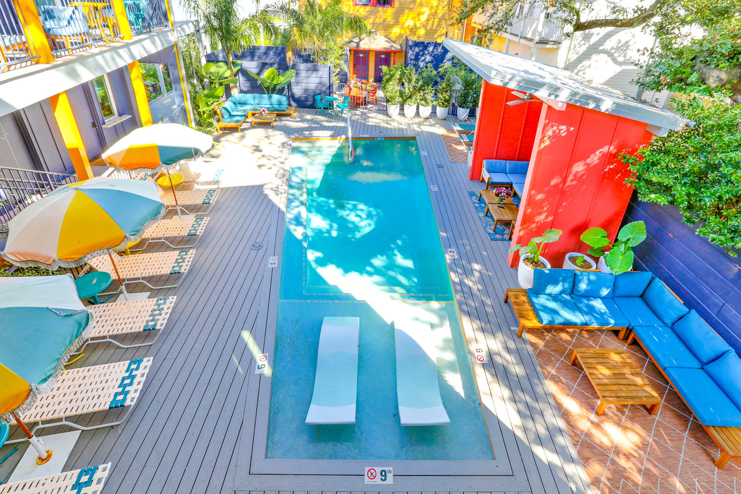 Vibrant courtyard pool at a retro boutique hotel — sunny outdoor plunge pool with two in-pool loungers, striped umbrellas, lounge chairs, and colorful seating areas