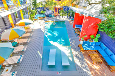 Vibrant courtyard pool at a retro boutique hotel — sunny outdoor plunge pool with two in-pool loungers, striped umbrellas, lounge chairs, and colorful seating areas