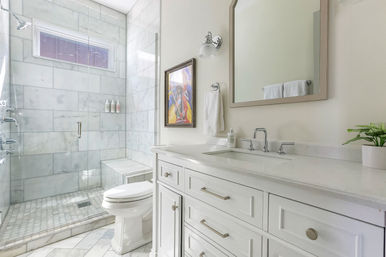 Bright spa-like modern bathroom with marble-tiled walk-in shower and bench behind frameless glass, white toilet, white shaker vanity with stone countertop and chrome faucet, mirror, towel and potted plant