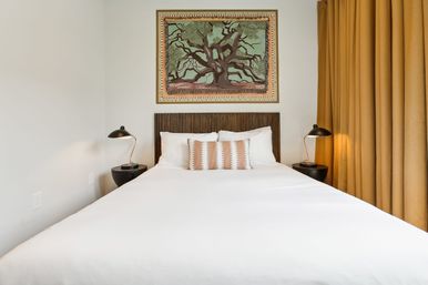 Bright, cozy minimalist bedroom interior with white duvet, dark wood slatted headboard, patterned lumbar pillow, framed tree artwork above, black bedside lamps and mustard-yellow curtains.