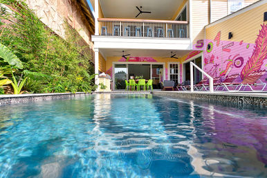 Low-angle view of a bright blue swimming pool leading to a two-story yellow house with balcony and covered patio, neon-green dining chairs, tropical plants and a pink Louisiana mural.