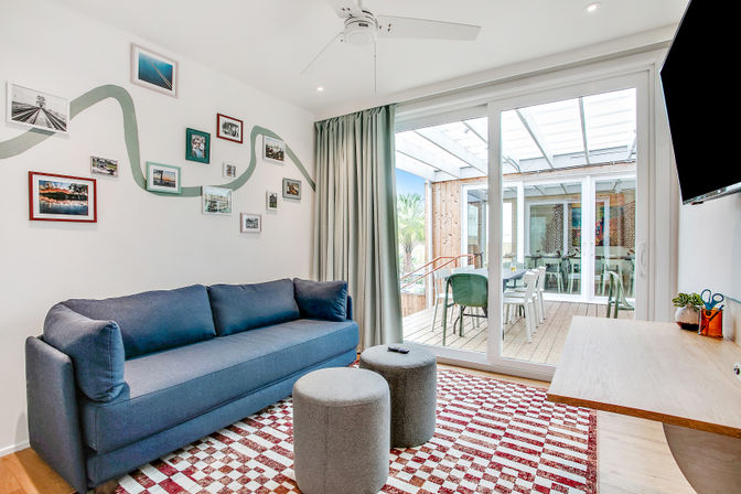 Bright contemporary living room with a navy sofa, gallery wall of framed photos, red-and-white check rug and two gray ottomans; sliding glass doors open to a covered outdoor dining patio, with wall-mounted TV, ceiling fan and light wood desk creating a sunny indoor-outdoor lounge.