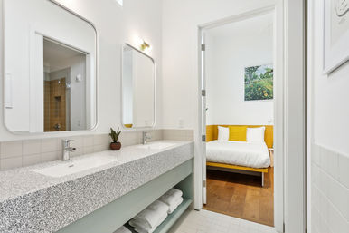 Bright modern bathroom with terrazzo double vanity, two rounded mirrors, chrome faucets, small potted plant, and view into adjacent white bedroom with yellow bed frame, yellow accent pillows and wood flooring.