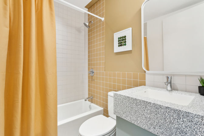 Cheerful modern bathroom with white tub-shower, mustard-yellow shower curtain and tiled accent wall, terrazzo-style speckled vanity countertop, wall mirror and small potted plant.