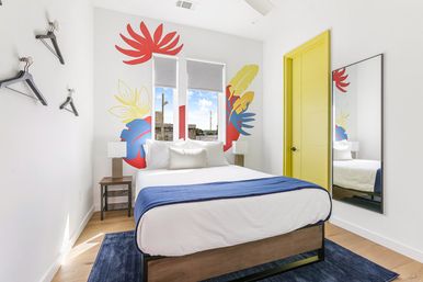 Sunlit modern bedroom with a colorful tropical wall mural behind a white bed dressed with a blue throw, yellow interior door, two tall windows overlooking urban rooftops, bedside tables and a full-length mirror.